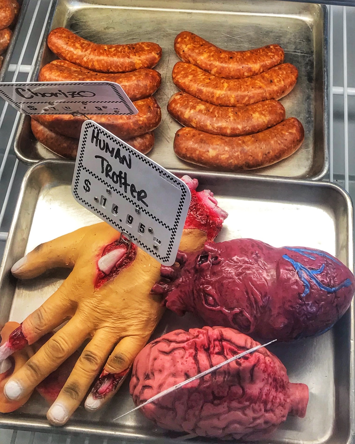 Figuras de plástico en forma de corazón, cerebro y mano a la venta en un mercado de Nueva Orleans con chorizo y otros alimentos reales al fondo, mostrando productos curiosos y gastronomía local