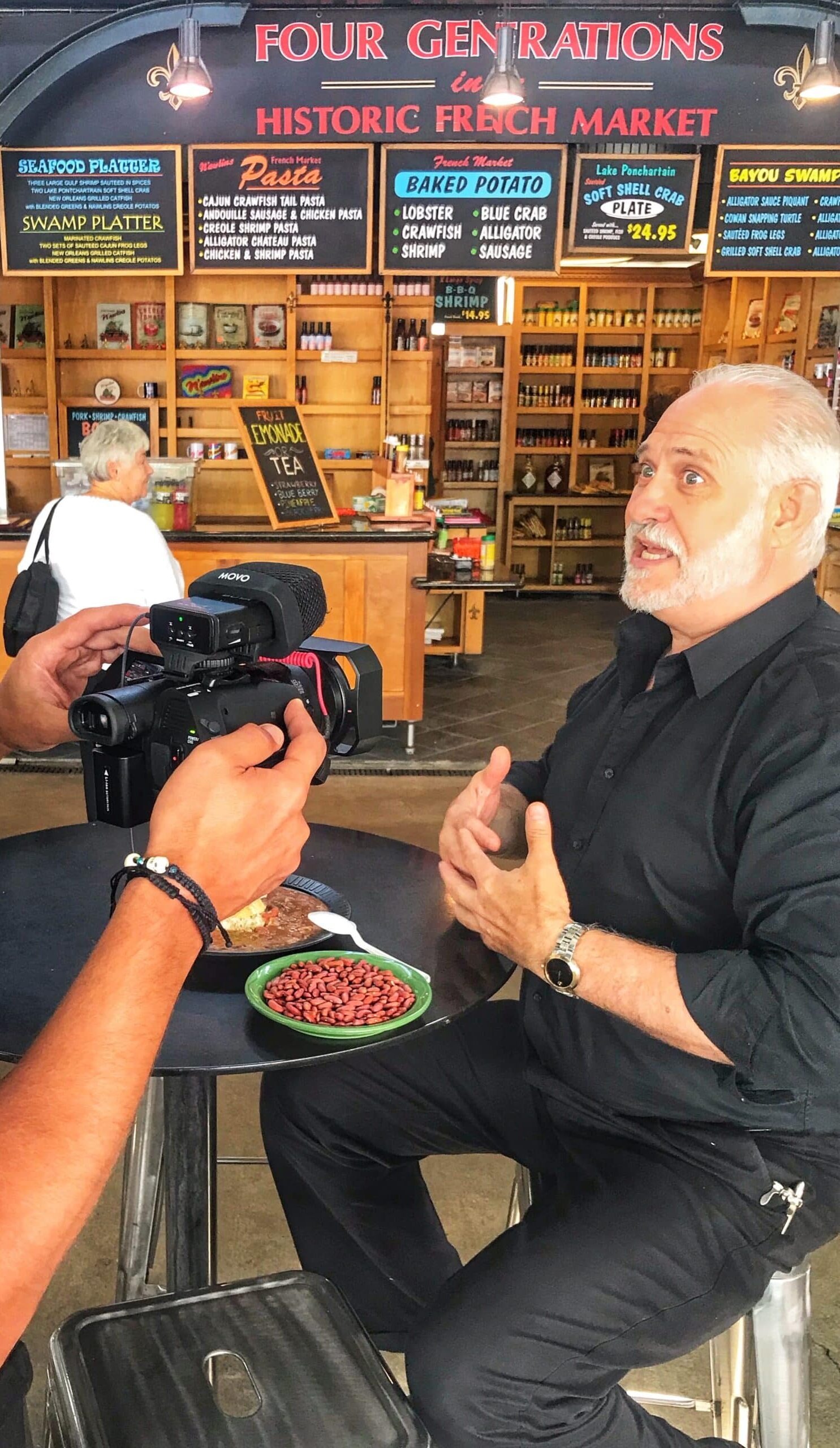 Hombre, en un Market de Nueva Orleans, siendo grabado mientras habla sobre productos y gastronomía local, mostrando el ambiente de mercado y la cultura culinaria de la ciudad