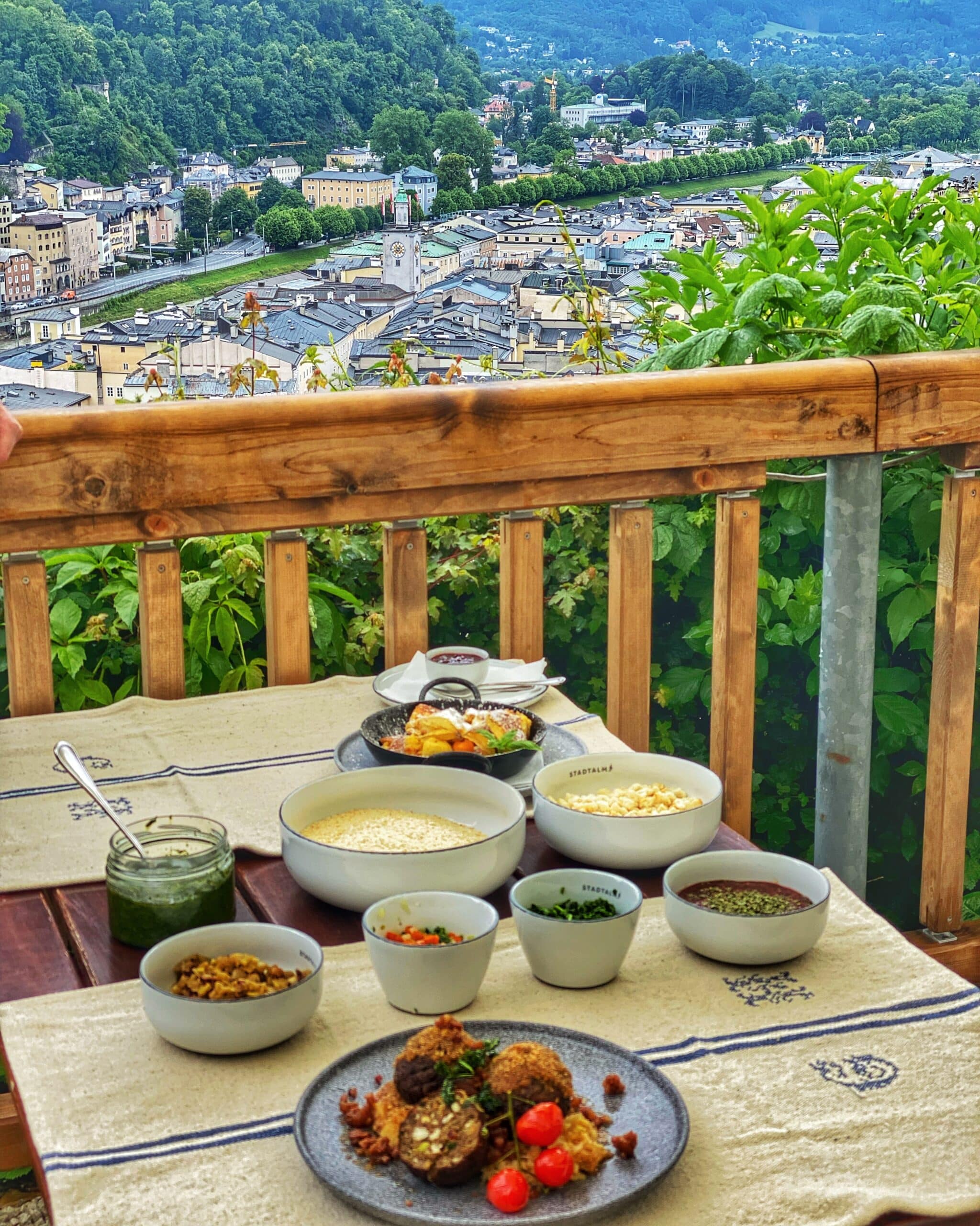 Variedad de platos tradicionales y servidos sobre una mesa con el paisaje urbano y montañoso de Salzburgo, al fondo, mostrando la gastronomía y el entorno de la ciudad.