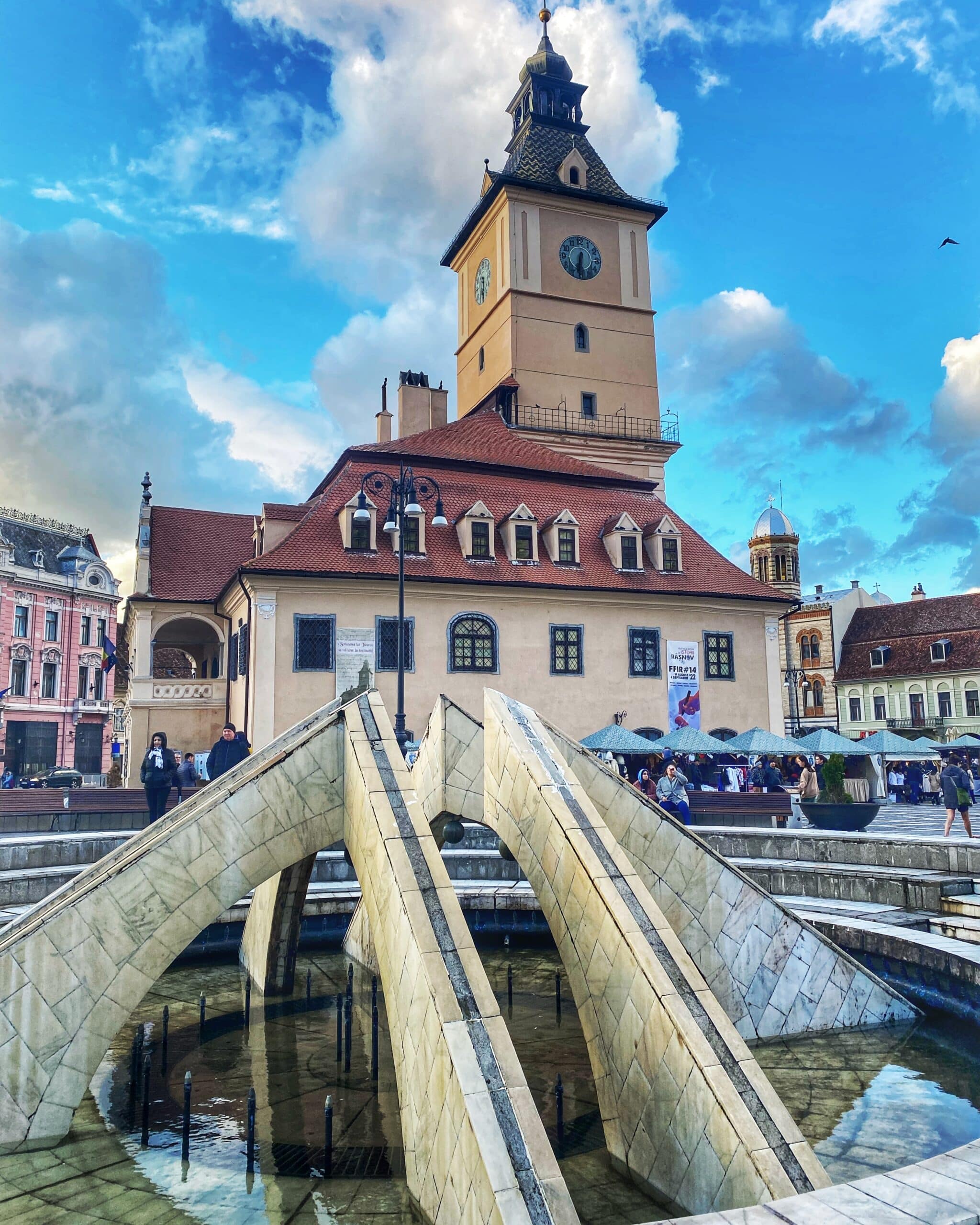 Vista de una plaza típica de Transilvania con una fuente en el centro y un edificio histórico de fondo, mostrando la arquitectura local y el ambiente urbano.