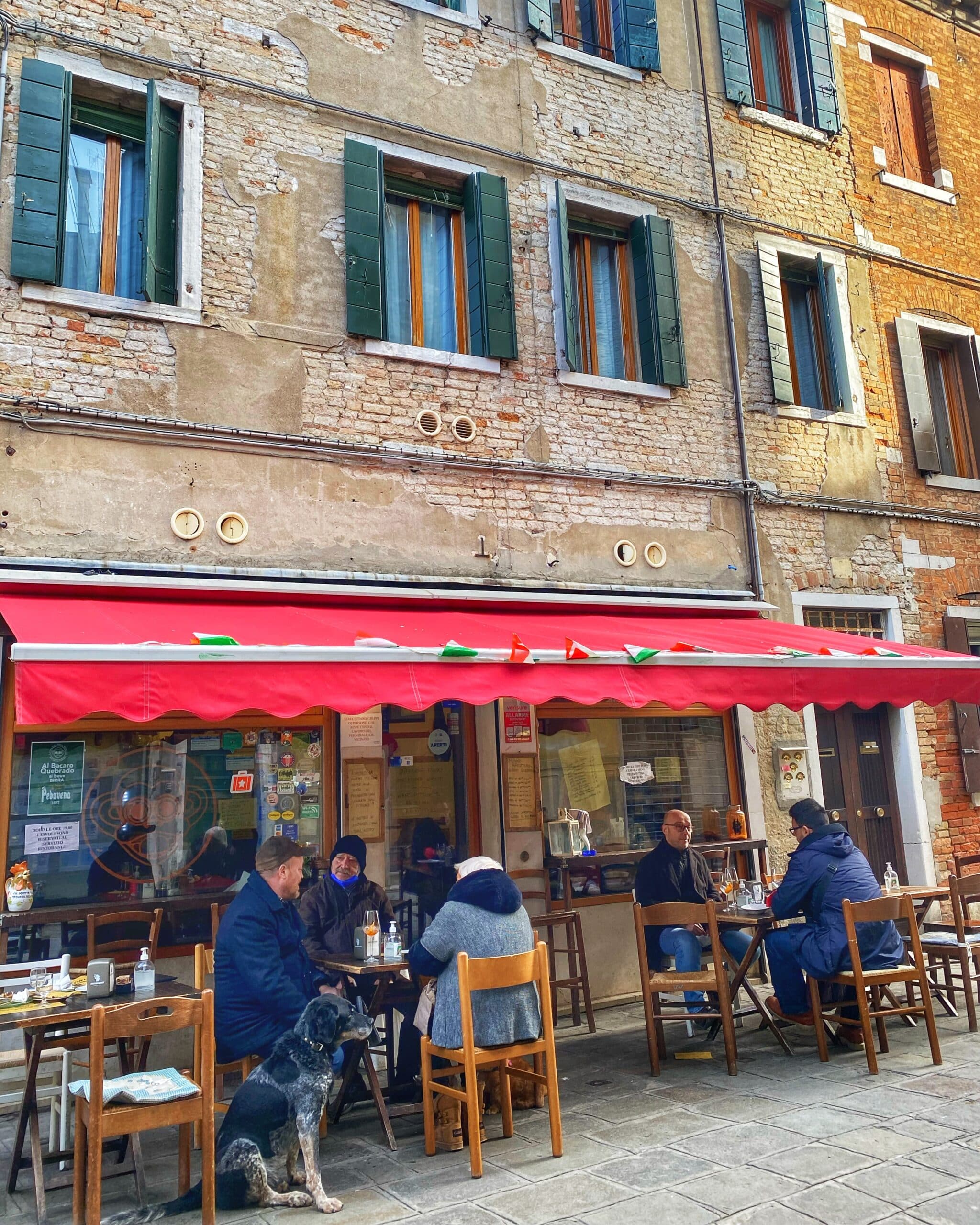 Personas sentadas, disfrutando en un bar tradicional de Venecia, con mesas al aire libre y la atmósfera característica de los canales italianos