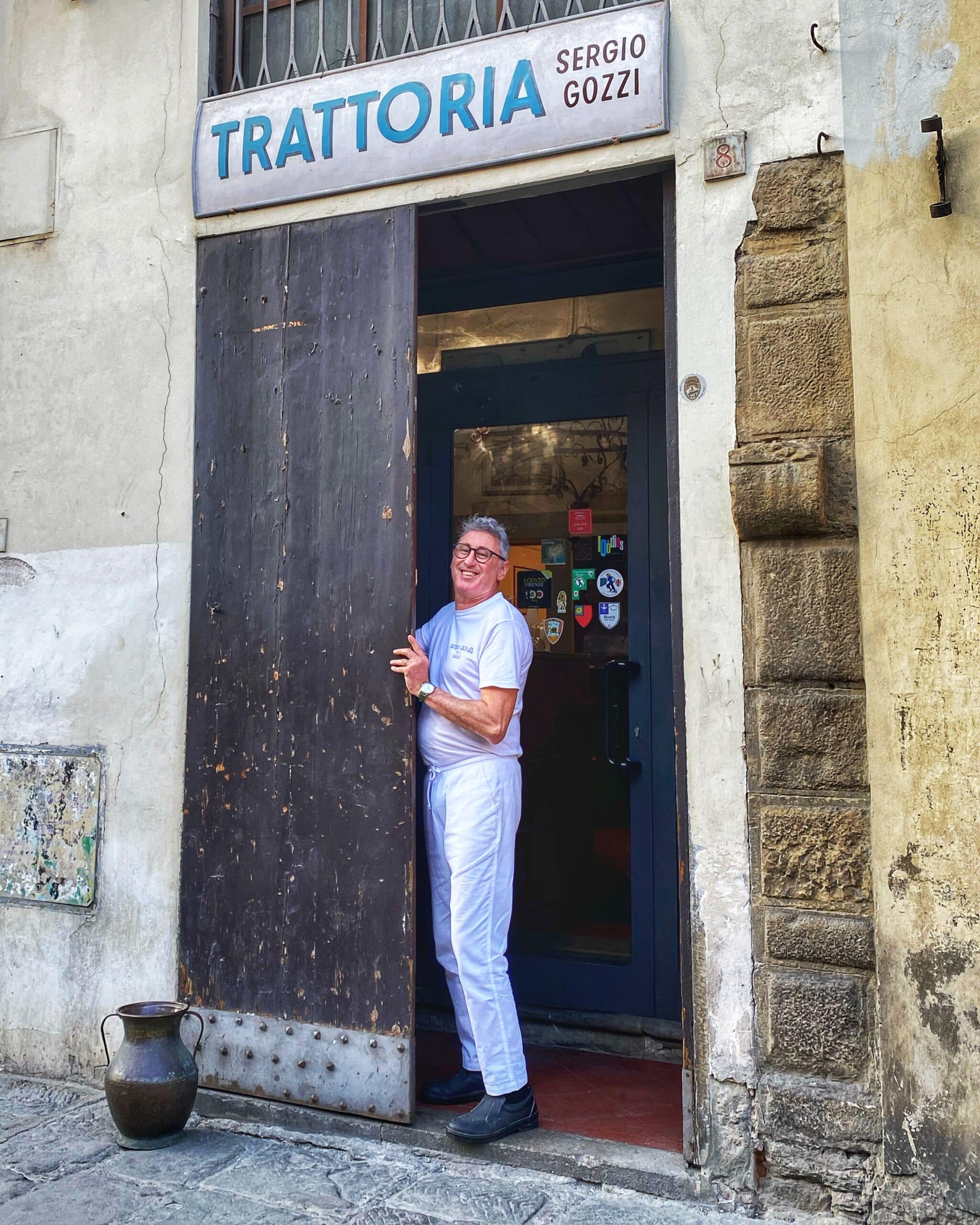 Hombre, sonriente, vestido de blanco, abriendo la puerta de su tienda en la toscana, mostrando la hospitalidad y el comercio local típico de la región.