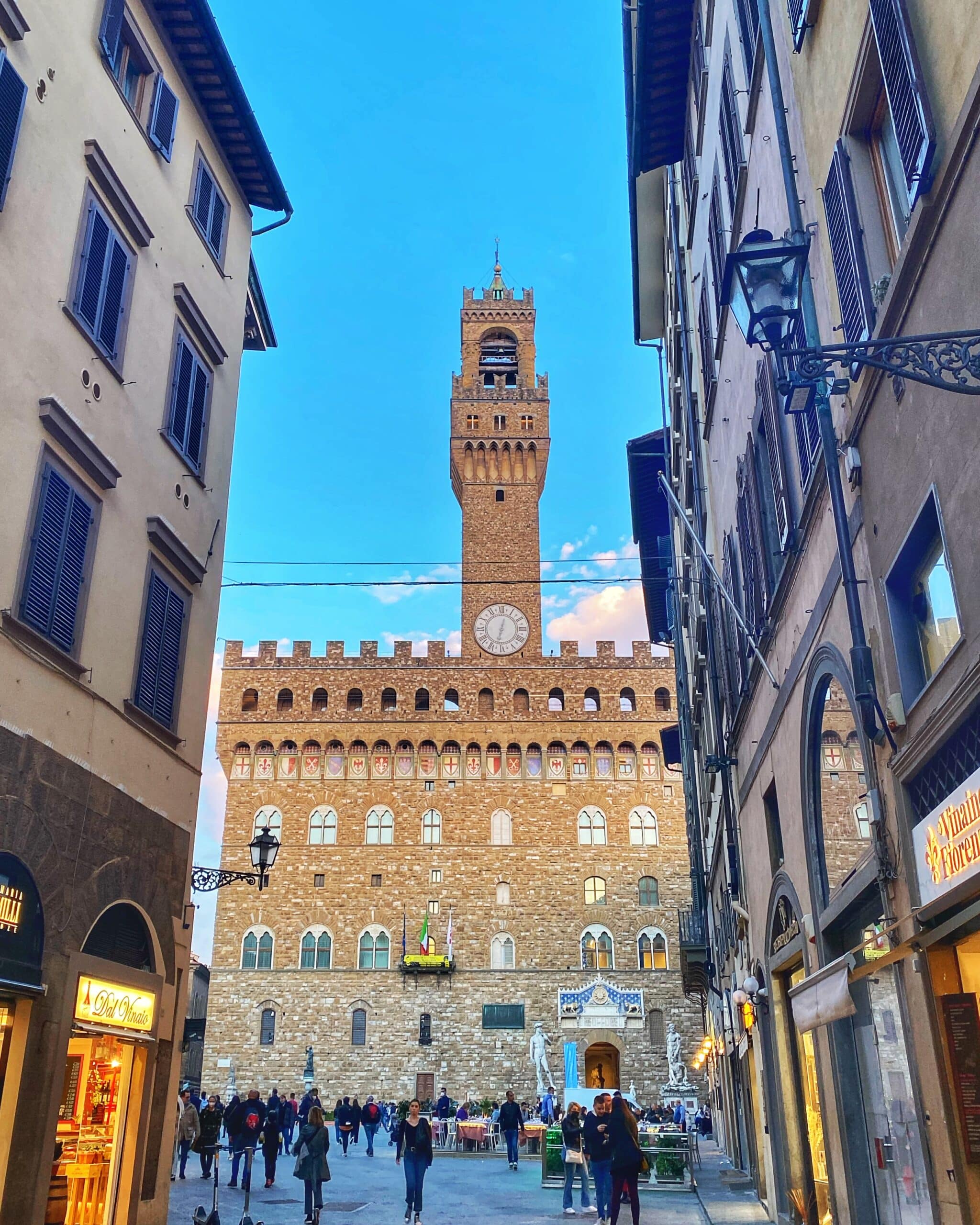 Personas caminando frente a un edificio histórico en la toscana, mostrando la arquitectura tradicional y la vida cotidiana de la región.