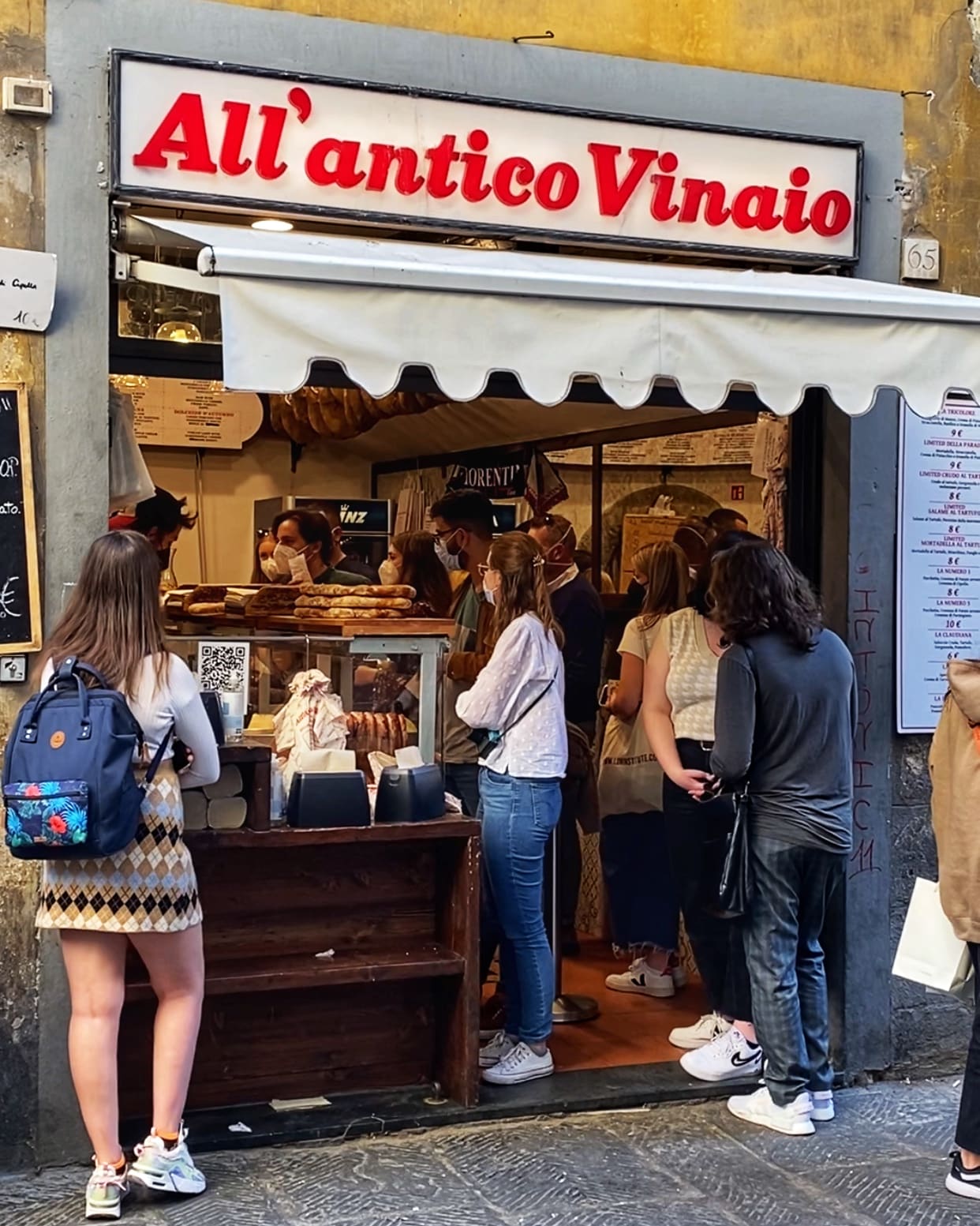 Personas haciendo cola frente a una tienda de la toscana, mostrando la vida cotidiana y la actividad comercial típica de la región.