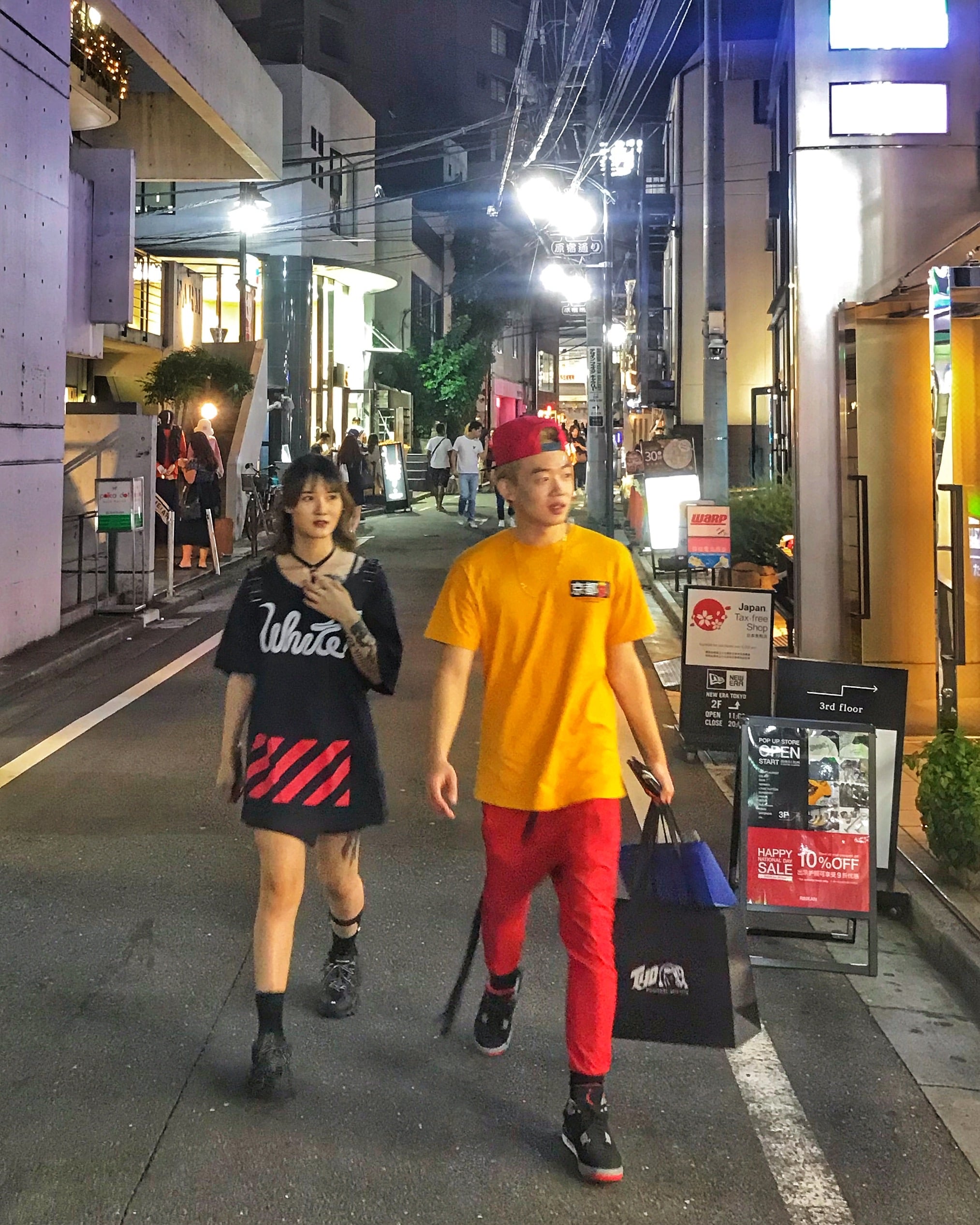 Calle iluminado de noche en Tokio, con dos personas caminando, una chica vestida de negro y un chico con ropa amarilla y roja, mostrando ambiente urbano nocturno japonés