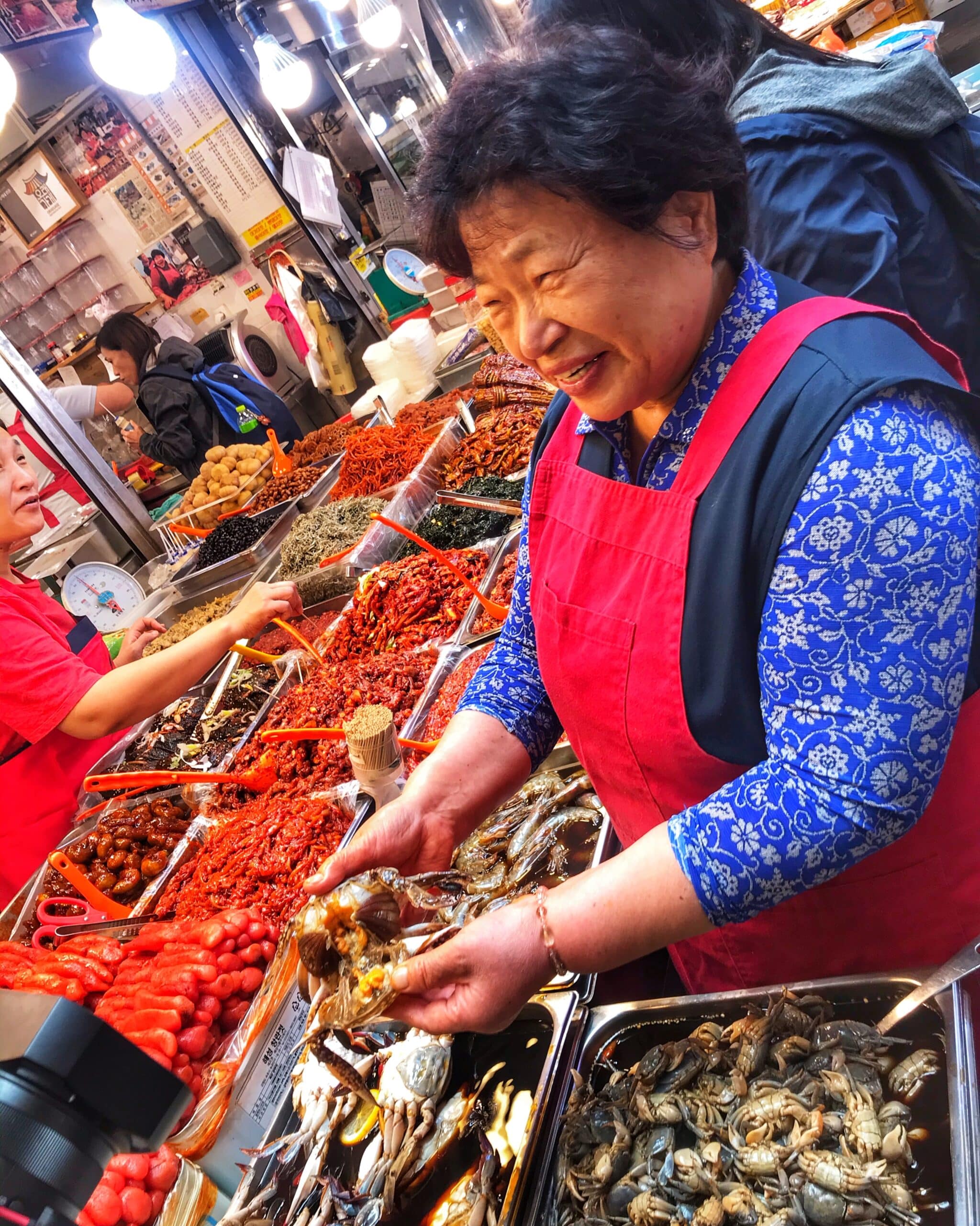 Mujer coreana, sonriente, siendo grabada en un mercado de seguros, rodeada de crustáceos y otros productos frescos, mostrando la gastronomía y ambiente animado del mercado coreano