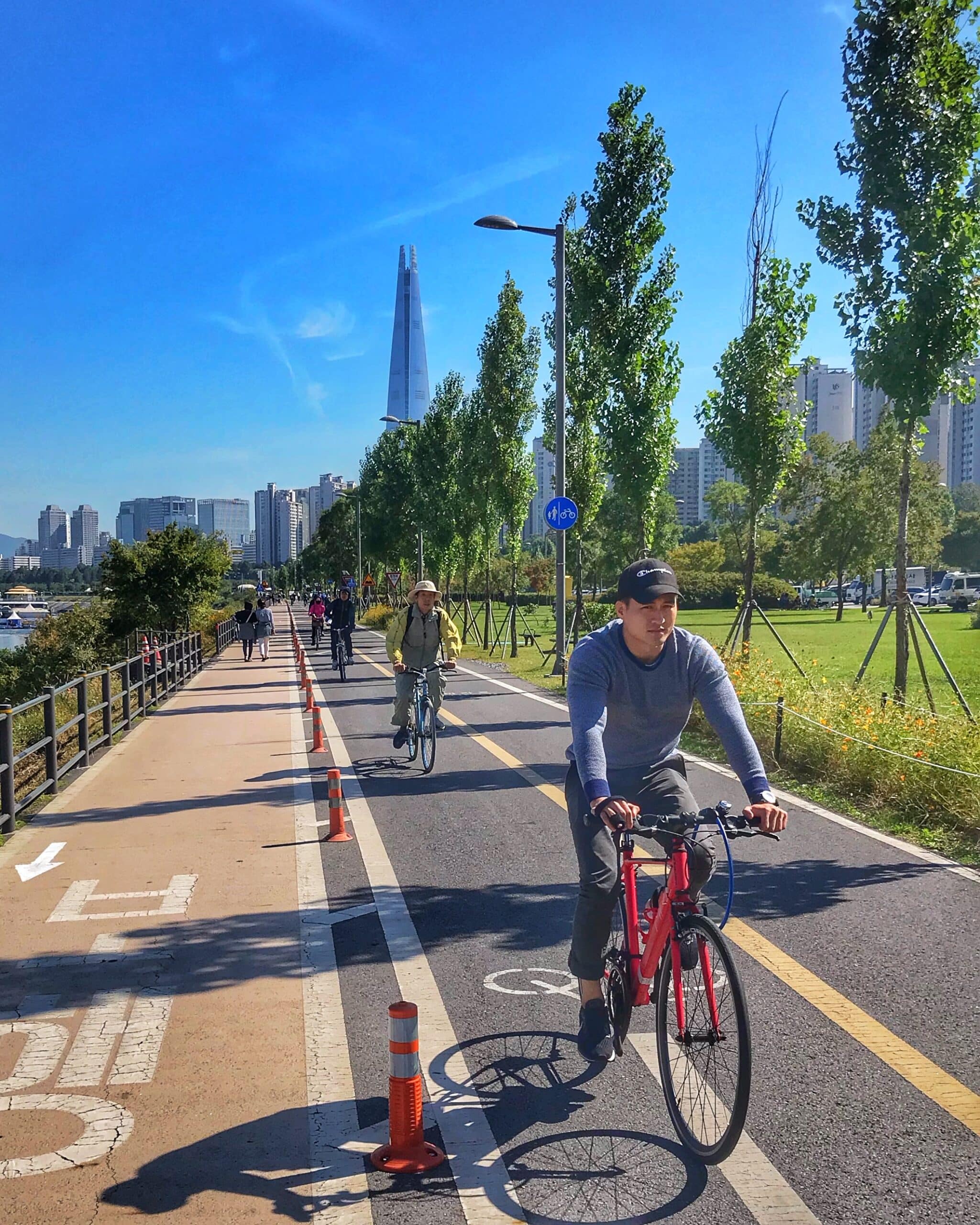 Parque en Seúl con personas caminando montando en bicicleta y disfrutando de actividades al aire libre, rodeado de áreas verdes y entorno urbano coreano